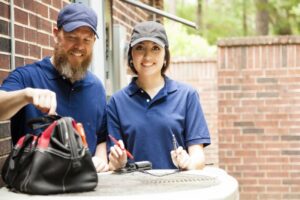 male-and-female-hvac-technicians-wearing-blue-polo-shirts-and-caps-outside-with-tool-box-on-ac-unit